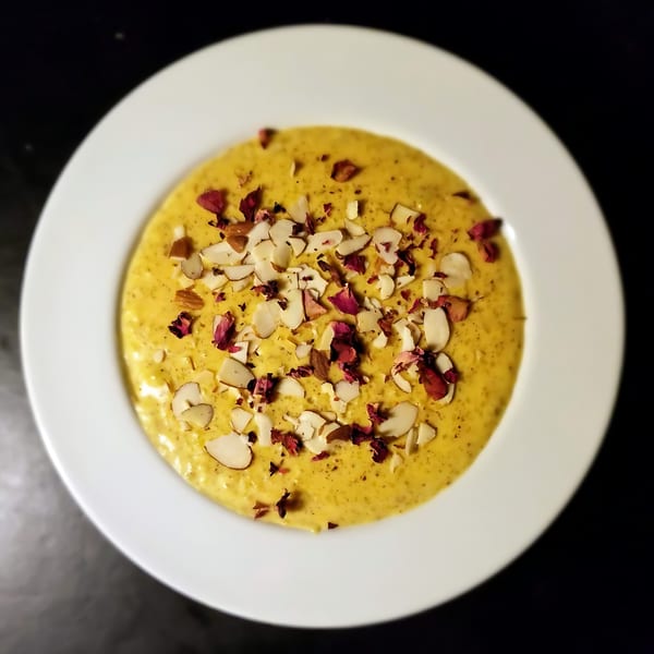 Halal Kheer, a creamy yellow dessert topped with sliced almonds and dried rose petals in a white bowl.